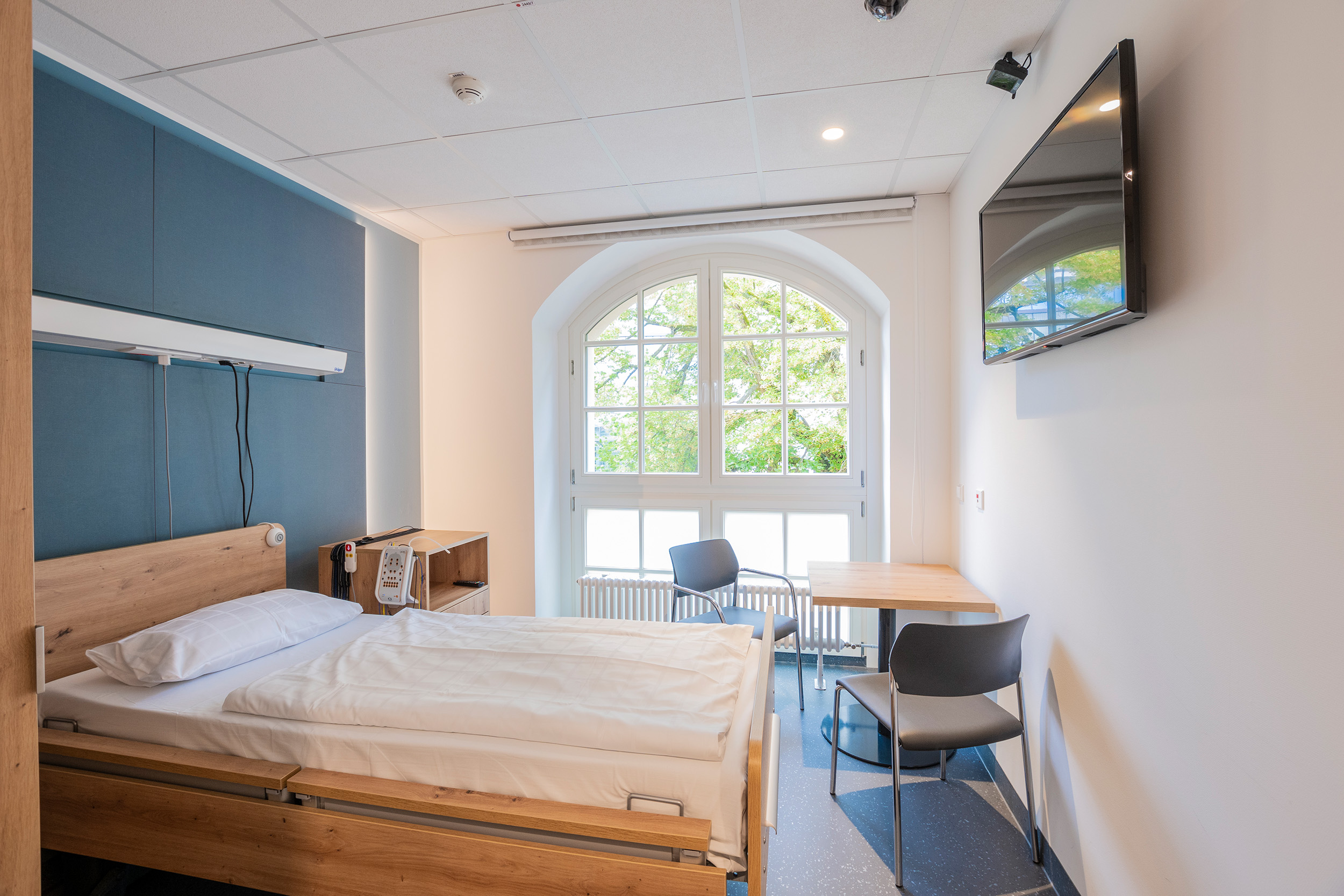 Patientenzimmer mit geräumigem Bett, großen Fenstern und Fernseher